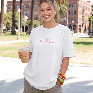 May include: A white short-sleeved t-shirt featuring the word pilates written in a pink cursive script across the center of the chest. The shirt has a crew neckline and a relaxed fit, made of a solid white fabric with no other patterns or designs.