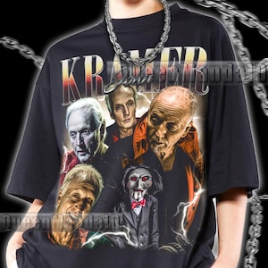 Könnte beinhalten: Schwarzes T-Shirt mit einem Grafikdesign im Vintage-Stil, das Charaktere aus dem Horrorfilm "Saw" und das Wort "Kramer" zeigt. Das Design beinhaltet eine Puppe mit roter Fliege und Blitzen.