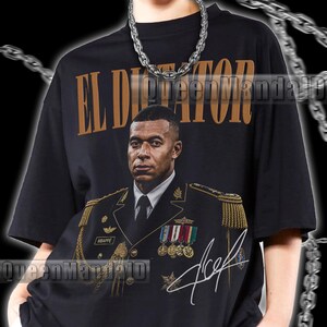 以下が含まれることがあります： 黒の半袖Tシャツで、軍服を着た人物のグラフィックプリントと、その上に金色の文字でEL DICTATORと書かれています。デザインにはMBAPPEと書かれたネームタグ、メダル、そして前面に白いサインのグラフィックが含まれています。
