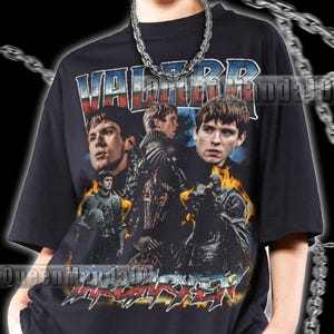 以下が含まれることがあります： 「VALOR」の文字と人物の画像が描かれたグラフィックデザインの黒いユニセックスTシャツ。クルーネックと半袖です。シルバーのチェーンネックレスが着用されています。