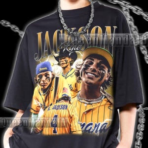 Peut inclure: Un t-shirt noir à manches courtes avec une impression graphique d'un joueur de baseball en uniforme rayé jaune. Le motif inclut le texte JACKSON et Kyle, avec plusieurs images du joueur portant une casquette et du maquillage noir sous les yeux. Un collier en chaîne argentée est porté sur le t-shirt.