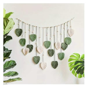May include: A macrame wall hanging featuring leaf shapes in shades of green and cream. The leaves are suspended from a horizontal string with wooden beads. The design is against a white wall, with green plants in the background.