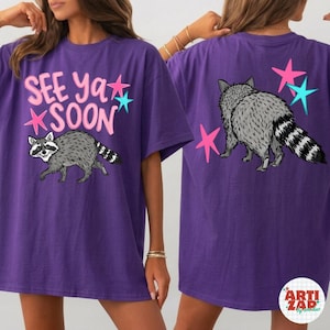 May include: Purple t-shirt with a raccoon graphic and the text "See Ya Soon". The design includes pink and teal stars. The back of the shirt features a raccoon and more stars.