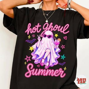 May include: A black t-shirt featuring a graphic of a ghost wearing pink sunglasses and platform sandals, holding a pink cocktail. The text reads hot Ghoul Summer in pink script, surrounded by colorful stars, hearts, and small purple bats.