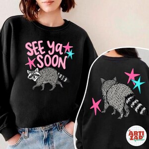 May include: Black sweatshirt featuring a raccoon illustration with the text "SEE YA SOON" in pink and blue. The back of the sweatshirt also has a raccoon illustration and pink and blue stars.