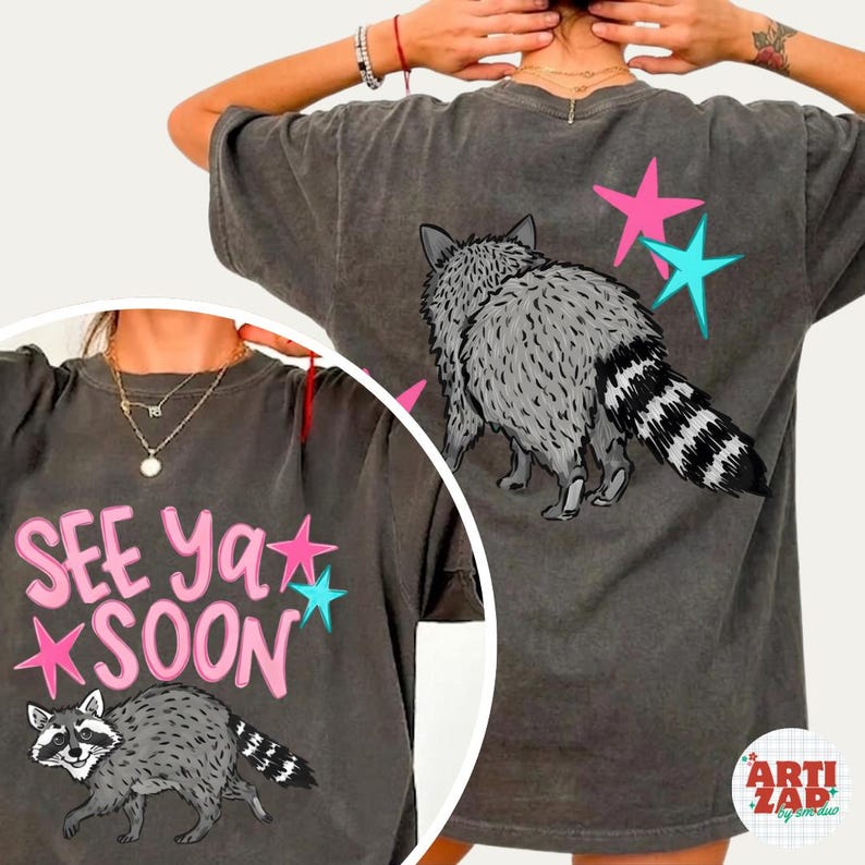 May include: Dark gray t-shirt featuring a raccoon illustration with a striped tail and the text "SEE YA SOON". Pink and turquoise stars accent the design. The back of the shirt also features a raccoon and stars.