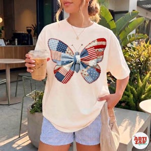Op de afbeelding: Crèmekleurig T-shirt met een groot vlinderontwerp gemaakt van patchworkpatronen in rood, wit en blauw, dat lijkt op de Amerikaanse vlag. De vlinder heeft een blauw-wit geruite strik. Het shirt wordt gecombineerd met blauw-wit gestreepte shorts.