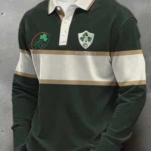 Puede incluir: Camiseta de rugby verde oscuro de manga larga con rayas blancas y beige. Presenta un cuello blanco, un escudo de trébol y el texto "Ireland Rugby" y "SINCE 1875".