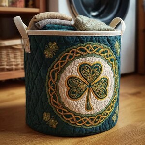 May include: A dark teal quilted laundry basket with a shamrock design. The basket has a central shamrock emblem surrounded by a gold Celtic knot design. It is filled with folded towels and has cream-colored handles.