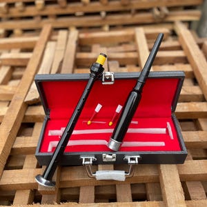 Puede incluir: Un juego de gaita negro con interior rojo, que contiene dos piezas de gaita negras, dos piezas pequeñas rojas y blancas y otros accesorios. Las piezas de la gaita son negras con detalles plateados y amarillos. El juego se muestra sobre una superficie de madera.