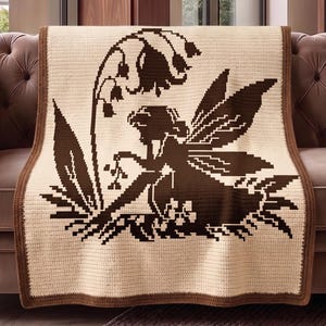 May include: A crocheted blanket featuring a fairy design in shades of brown and cream. The fairy is depicted with wings, sitting near a flower. The blanket has a brown border and is draped over a brown sofa.