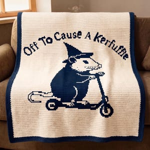May include: A cream-colored crocheted blanket with a navy blue border. The blanket features a navy blue illustration of an opossum riding a scooter, wearing a witch hat, and the text "Off To Cause A Kerfuffle".
