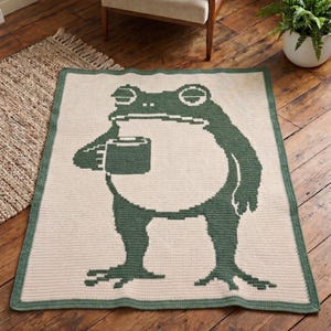 Puede incluir: Una manta color crema con un borde verde y un gran diseño de rana verde. La rana sostiene una taza. La manta es rectangular y parece estar hecha a ganchillo.