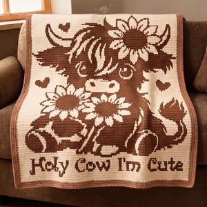Puede incluir: Una manta de ganchillo con un diseño de vaca de las Tierras Altas marrón, girasoles y corazones sobre un fondo crema. La manta tiene un borde marrón y el texto "Holy Cow I'm Cute". La manta está sobre un sofá marrón.