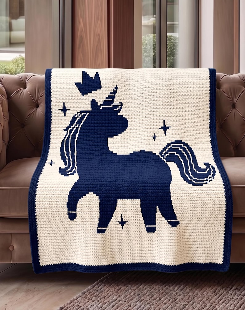 Pu&ograve; includere: Una coperta all'uncinetto color crema con un design a unicorno blu navy. La coperta presenta un bordo blu navy, una silhouette di unicorno, una corona e accenti di stelle. La coperta &egrave; drappeggiata su un divano marrone.
