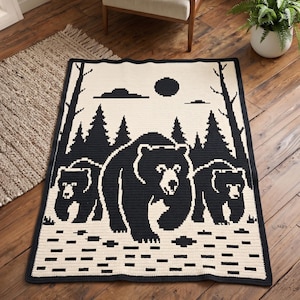 May include: A rectangular crochet blanket featuring a black and cream pixelated design of a large bear walking with two smaller bears in a forest setting. The scene includes tall pine trees, a sun or moon, and clouds against a light background.