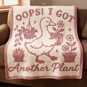 May include: A cream-coloured crocheted blanket with a pink border and design. The design features a duck carrying potted plants, with the text "OOPS! I GOT Another Plant". The blanket is folded over a brown sofa.