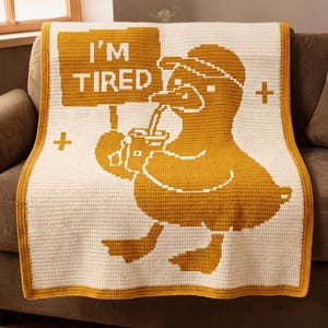 May include: A crocheted blanket featuring a large yellow duck holding a sign that reads "I'M TIRED" and a drink. The blanket has a white background with yellow accents and a yellow border. The duck is wearing a hat and drinking from a cup.