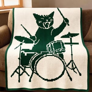 May include: A rectangular cream-colored throw blanket featuring a dark green pixelated graphic of a cat playing a drum set. The cat is depicted with drumsticks raised, sitting behind a bass drum, snare, and cymbals, all bordered by a dark green edge.