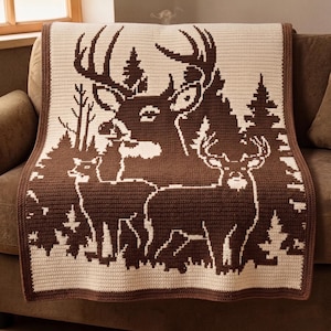 May include: A crocheted blanket featuring a brown and cream deer scene. The blanket displays a buck with large antlers, two smaller deer, and stylized trees. The border is a solid brown, and the blanket is draped over a brown sofa.