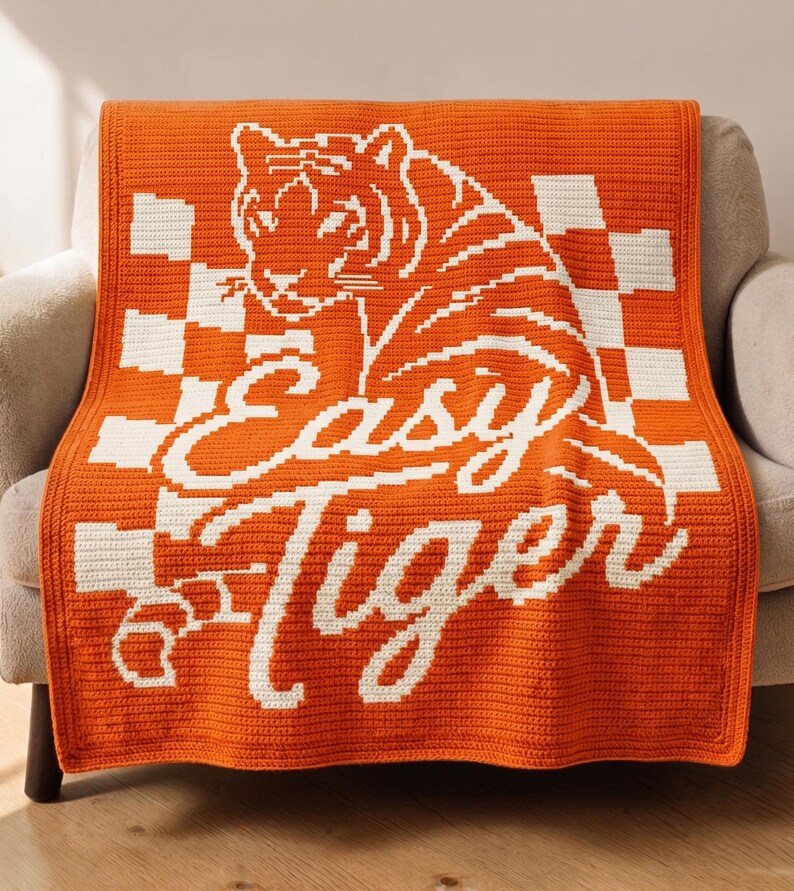 Pu&ograve; includere: Una coperta arancione all'uncinetto con un disegno di tigre bianca e le parole "Easy Tiger". La coperta presenta un motivo a scacchi ed &egrave; drappeggiata su una poltrona di colore neutro. La coperta &egrave; realizzata in un materiale morbido e strutturato.