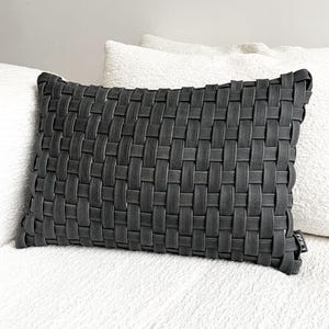 May include: A rectangular charcoal cushion with a woven design. The surface is made of interwoven strips, creating a textured pattern. The cushion is placed on a white surface, with other white cushions in the background.
