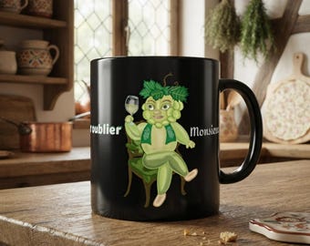 Peut inclure: Un mug en céramique noire illustré d'un personnage en forme de grappe de raisin verte, vêtu d'un gilet vert, assis sur une chaise et tenant un verre de vin. Le texte "troublier" et "Monsieur" est imprimé en blanc de chaque côté du personnage.