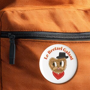 Peut inclure: Un bouton blanc avec le texte "Le Bretzel Galant" en rouge, représentant un personnage de bretzel de dessin animé portant un chapeau noir et une écharpe rouge en forme de cœur. Le bouton est sur un sac à dos orange.