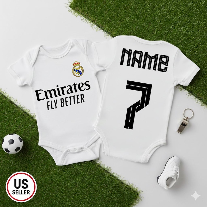 May include: Two white baby onesies with black text and graphics. One has "Emirates FLY BETTER" and a team logo. The other has "NAME" and the number "7". A soccer ball, whistle, and baby shoe are also in the image.