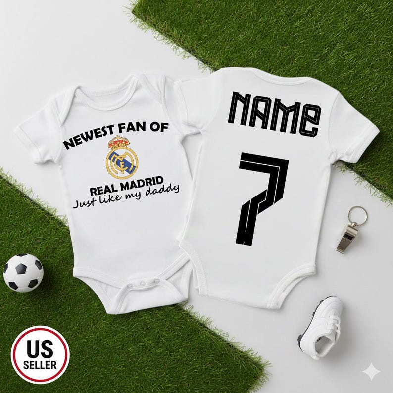 May include: Two white baby onesies with soccer-themed designs. One features the text "NEWEST FAN OF REAL MADRID" and a team logo. The other has "NAME" and the number 7. A soccer ball, whistle, and shoe are also present.