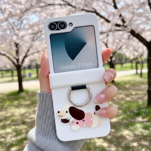 Puede incluir: Funda blanca para teléfono Galaxy Z Flip6 con diseño de perro de dibujos animados. La funda tiene un soporte de anillo plateado y la pantalla del teléfono muestra el texto "Galaxy Z Flip6".