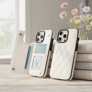 Puede incluir: Dos fundas blancas para iPhone con bordes negros. Una funda está abierta, mostrando ranuras para tarjetas de color azul claro. La otra funda tiene un patrón acolchado. Un jarrón de flores está en el fondo.