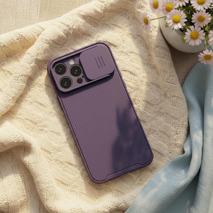 May include: A purple phone case with a camera cover is displayed on a cream-colored knitted blanket. The case has a textured back and a sliding cover for the camera lenses. A small vase of daisies is in the background.