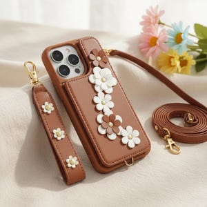 May include: A brown leather phone case with a strap and wristlet, decorated with white and brown flower details. The case includes a card slot and gold-coloured hardware. The wristlet and strap also have flower accents.