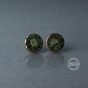 Può includere: Due orecchini a bottone rotondi con un centro strutturato verde scuro e un bordo color bronzo. Gli orecchini hanno un bordo bianco e sono su uno sfondo grigio. Un accessorio unico.