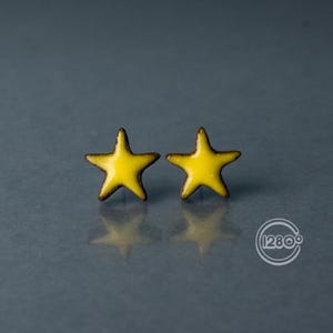 May include: A pair of yellow star-shaped stud earrings with a dark brown border. The earrings are set against a grey background, with a reflection visible below. The earrings are a jewelry item.