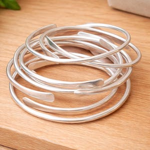 Puede incluir: Un conjunto de pulseras plateadas, abiertas y apiladas. Las pulseras tienen un acabado liso y pulido, con un diseño sencillo y elegante. Las pulseras están colocadas sobre una superficie de madera marrón claro.