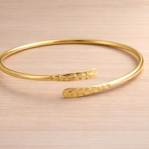 May include: A gold-colored open cuff bracelet. The bracelet has a hammered texture on the ends and a smooth, rounded band. The jewelry is displayed on a light-colored wooden surface.