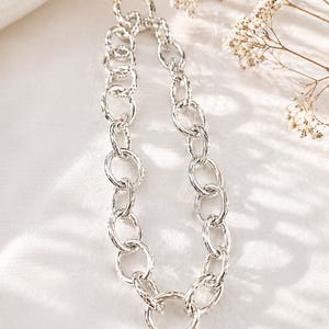 Puede incluir: Un collar de cadena plateada con eslabones ovalados texturizados y un gran cierre circular. El collar se muestra sobre una superficie blanca con iluminación suave, un producto de joyería.