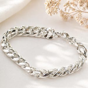 Puede incluir: Un brazalete de cadena de plata. El brazalete, hecho de eslabones entrelazados, presenta un cierre de mosquetón. La joya se exhibe sobre una superficie blanca, con flores secas en el fondo.