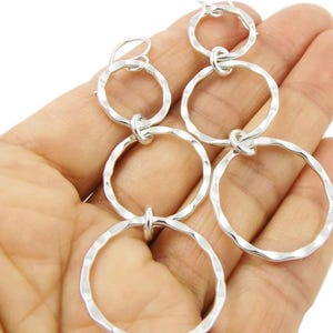 May include: Silver-toned dangle earrings featuring a series of connected hammered rings. The earrings have a textured surface and are suspended from a simple hook. The design is modern and minimalist, suitable for everyday wear or special occasions.