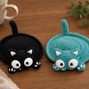 May include: Two round crochet coasters shaped like cats, one in black and one in teal. Each coaster features a cat head with large white eyes, black pupils, white inner ears, and white paw details. A small tail extends from the top of each circular body.