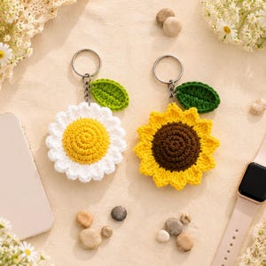 May include: Two crochet flower keychains featuring a silver metal ring. One keychain has white petals with a yellow center, and the other has yellow petals with a brown center. Each flower includes a small green leaf attached to the metal chain.