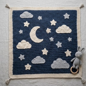 Peut inclure: Une couverture de bébé au crochet bleu marine avec un thème de ciel nocturne. Elle présente des nuages, des étoiles et un croissant de lune blancs et gris. Un hochet lapin gris avec un anneau en bois est à droite. Bordure et pompons crème.