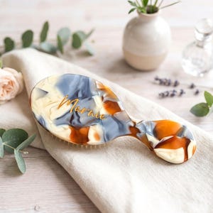 Op de afbeelding: Een ovale haarborstel met een handvat, met een gemarmerd ontwerp in crème, blauw en oranje tinten. De naam "Marnie" is in gouden letters gegraveerd. De borstel ligt op een crèmekleurige doek, met bloemaccenten.
