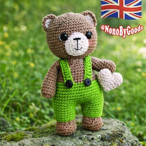 Op de afbeelding: Een handgemaakte gehaakte teddybeer met bruine vacht, witte accenten en een groene tuinbroek. De beer houdt een klein, gebroken wit hart vast. De tekst "@NonoByGoods" is zichtbaar.