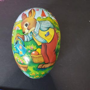 May include: An egg-shaped tin container with a colorful illustration of a rabbit watering flowers in a garden. The rabbit wears a yellow vest and red pants. A basket of fruit sits nearby.