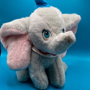 May include: A plush Dumbo the Elephant toy. The elephant is primarily gray with pink inner ears and blue eyes. It has a blue bow on its head and a white collar with a red trim. The toy is sitting on a blue background.