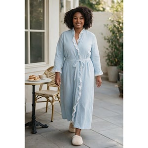 May include: A light blue, long-sleeved robe featuring a subtle white pattern, a ruffled white lace trim along the front opening and cuffs, and a matching fabric tie belt at the waist. The garment reaches mid-calf and has a stand-up collar.