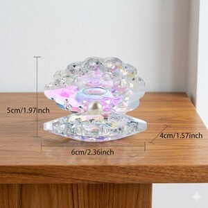Pode incluir: Concha de cristal iridescente com uma p&eacute;rola no centro. A concha decorativa tem 6 cm de largura, 5 cm de altura e 4 cm de profundidade. A concha est&aacute; sobre uma superf&iacute;cie de madeira.